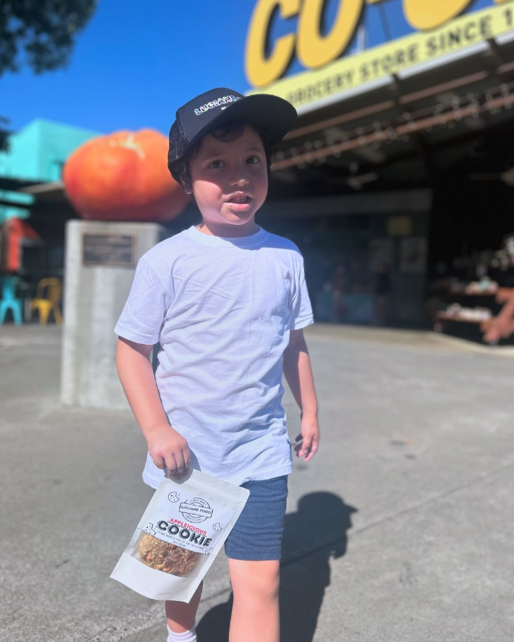 Lucian wearing a Lucian's Food cap holding an Applelicious Cookie pouch outside the Davis Food Co-Op